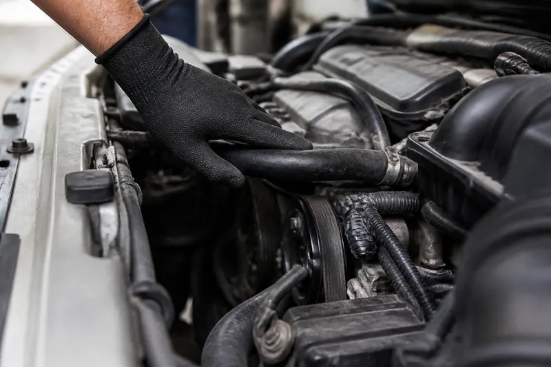 A gloved hand checking a radiator hose and drive belt in an engine bay